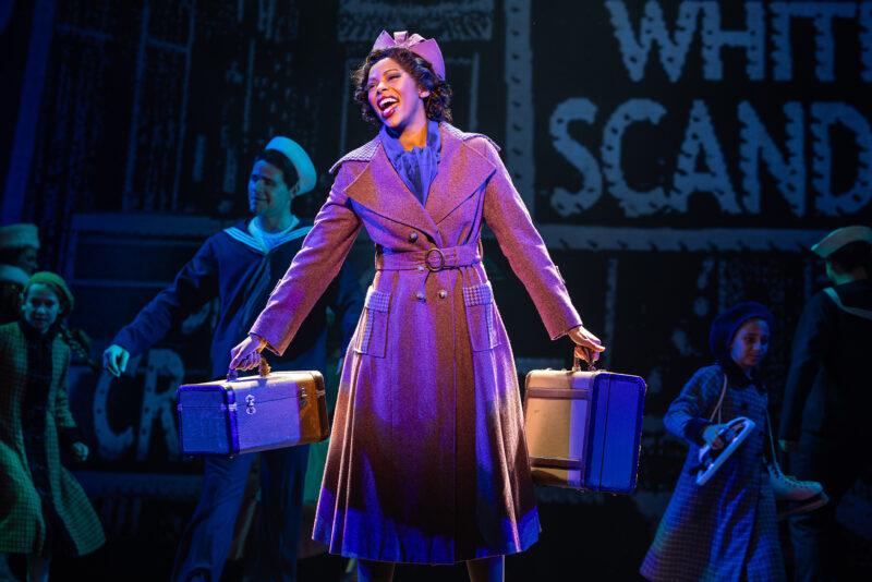 Savannah Fisher in the North American Tour of Annie. A woman, wearing a purple trench coat and matching hat, holds luggage in both hands as she sings. People walk casually behind her.