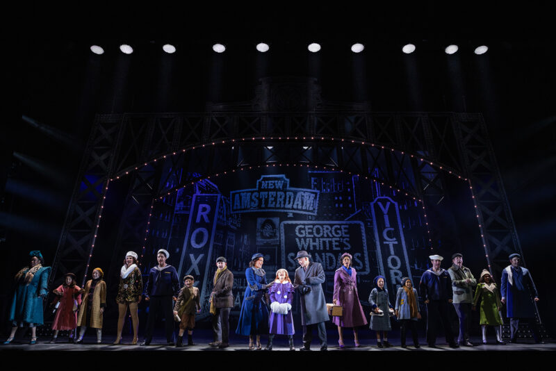 The cast of the North American Tour of Annie. A polished Annie stands center looking hopefully in the distance with her new family beside her. Onlookers of various societal statuses surround them. The backdrop reflects a vintage Times Square.