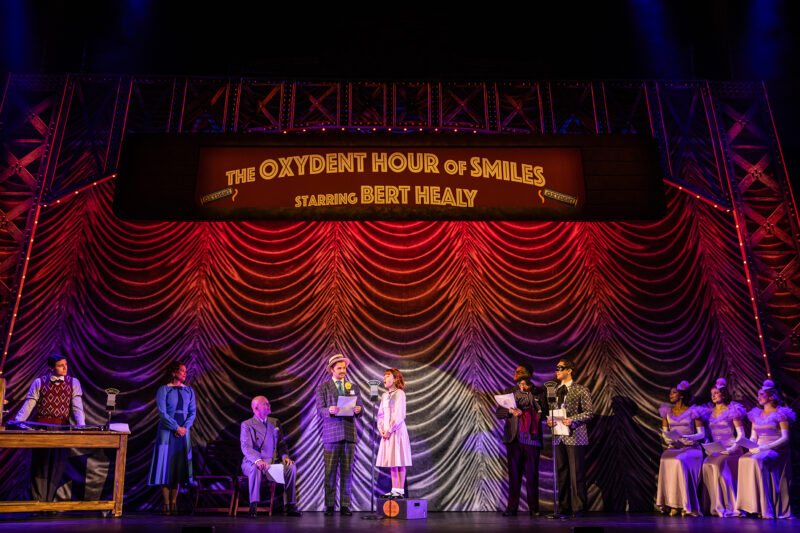 The cast of the North American Tour of Annie. Annie stands on a soapbox as she sings into a micrphone, a group of people around her watching on. A large sign reading “The Oxydent Hour of Smiles Starring Bert Healy” hangs above them.