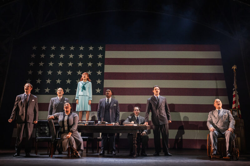 The cast of the North American Tour of Annie. Annie, wearing a mint green gown and jacket, stands on a desk. A group of sharply dressed individuals surround Annie. All face the audience singing. The backdrop is a large American flag.